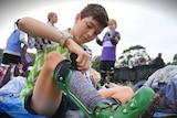 A young boy in a soccer jersey is putting on a pair of green boots that strap around his calf.