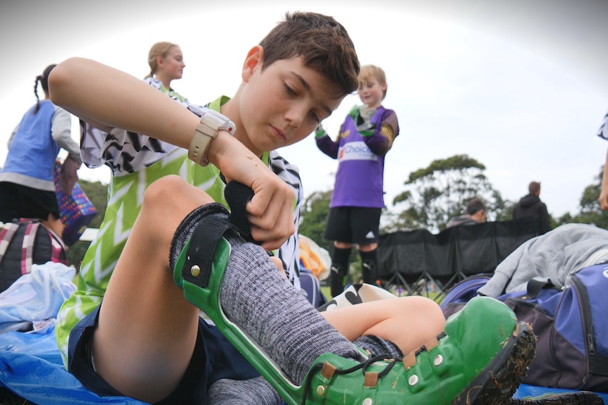 A young boy in a soccer jersey is putting on a pair of green boots that strap around his calf.