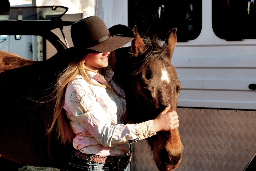 A girl wearing a wide brimmed hat pats a horse.