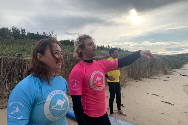 Three people stand on the beach in different colored overalls and shirts pointing to the water