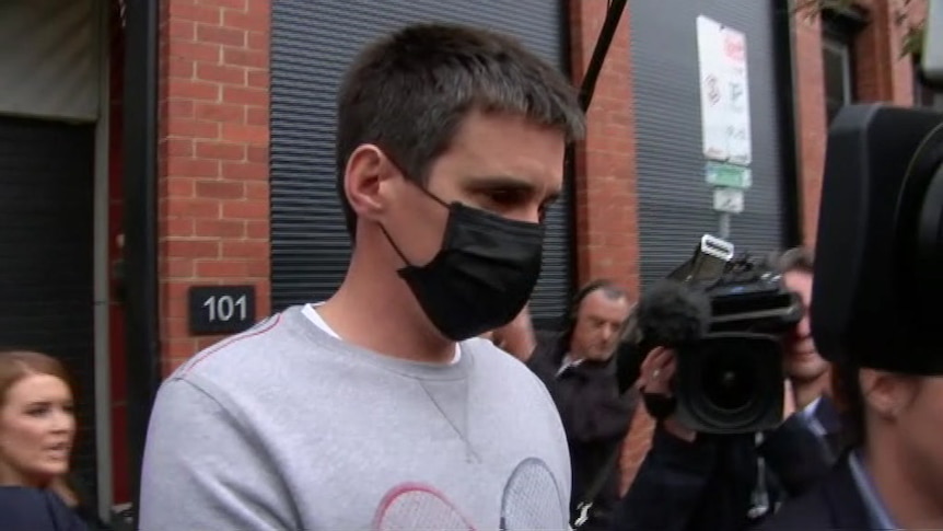 A man wearing a face mask is escorted by police, surrounded by a media scrum.