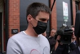 A man wearing a face mask is escorted by police, surrounded by a media scrum.