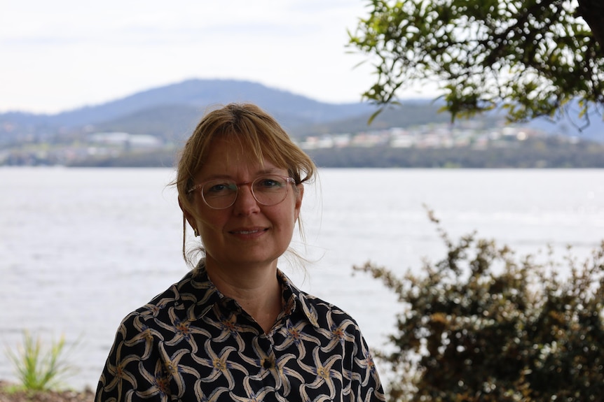 A woman standing near a river with hills and out of focus houses in the background