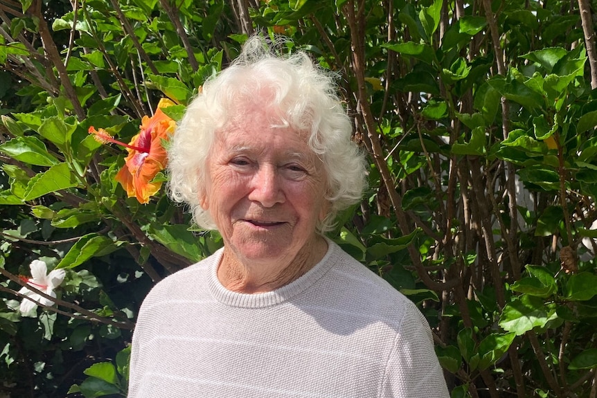 91-year-old woman with white hair standing in front of a bush with a smile on her face.