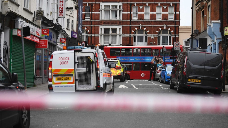 A London street with a red double decker bus inside a police tape with a police dog vehicle parked.