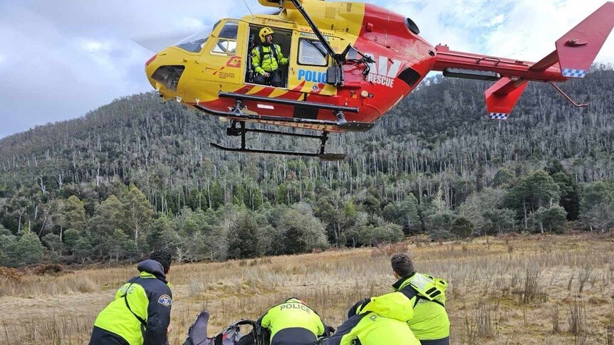 A rescue helicopter hovers a rescue party who are wearing hi-vis, they are on grass in the tasmanian wilderness.