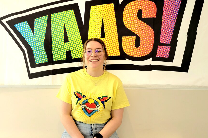 A woman in a yellow shirt sits in front of a multi-coloured sign that says YAAS!