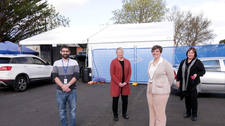 Four people stand 1.5 metres apart smiling. They stand in front of a white marquee outside