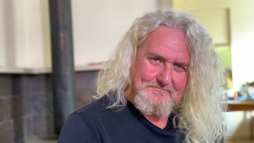 A man with long grey hair and a beard sits at a table and looks at the camera.