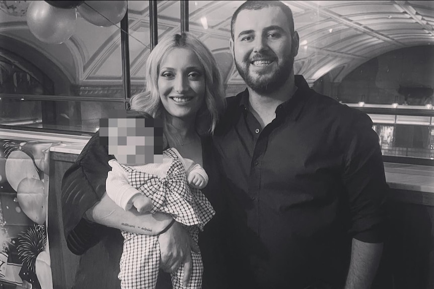 A couple smiling in a black and white photo, holding a baby.