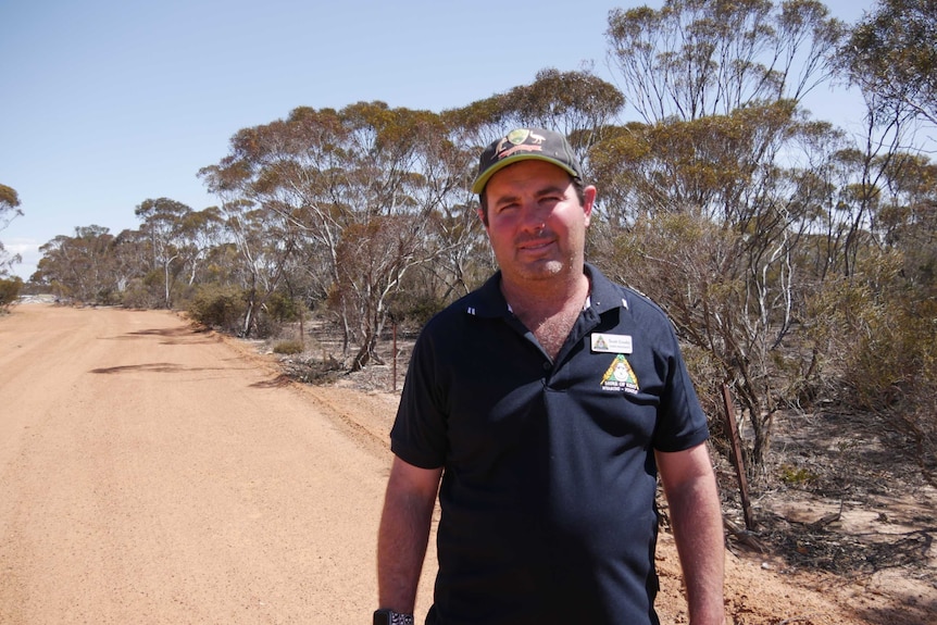 A man in a baseball cap and polo shirt stands on a dusty outback road.