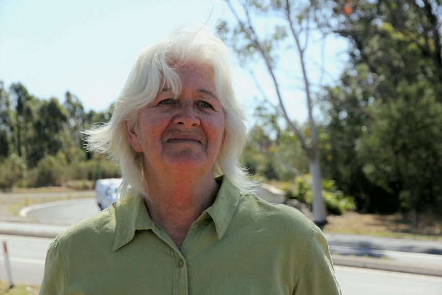 Woman with white hair in green shirt with resigned and upset expression stands in front of highway backdrop 