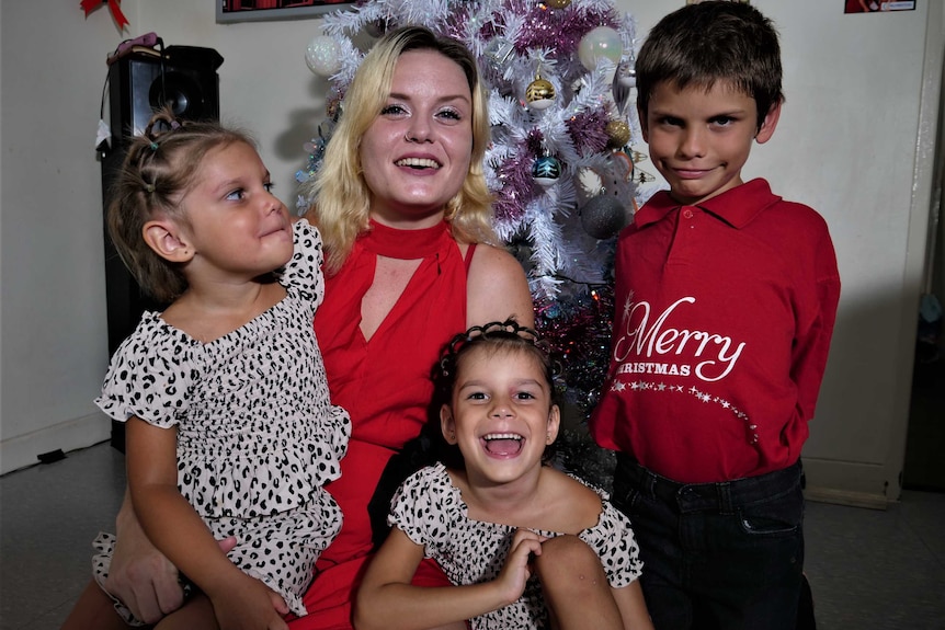 Mum in red dress with three kids kneeling in front of the home Christmas tree. All happy.