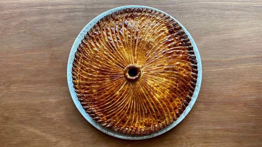 A intricately carved golden pastry pie in a wave pattern set on a bone-coloured ceramic plate on a wooden table.