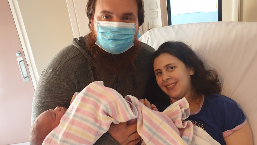 A woman sits up in a hospital bed with a man next to her cradling a swaddled newborn baby.