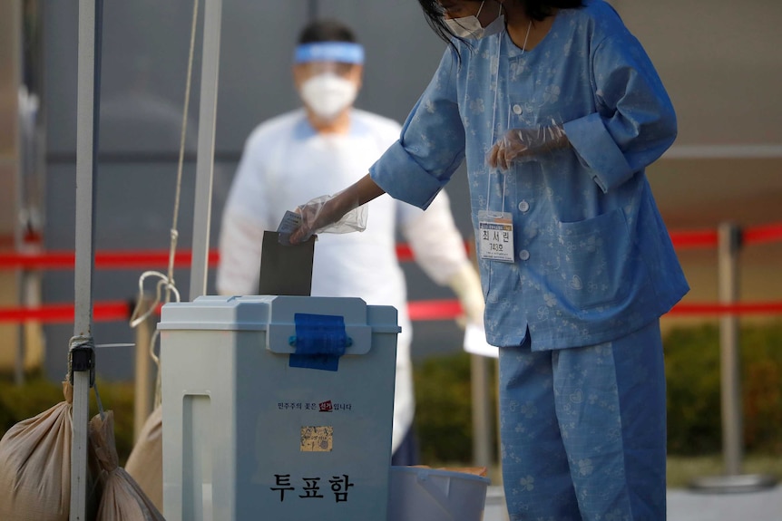 A woman wearing a mask and blue pyjamas casts vote in front of man in hazmat suit.