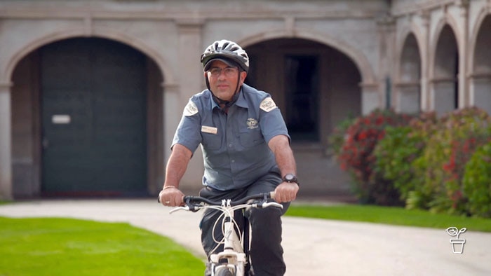 Man in grey shirt and helmet riding bicycle through the grounds of a large building courtyard with formal garden.