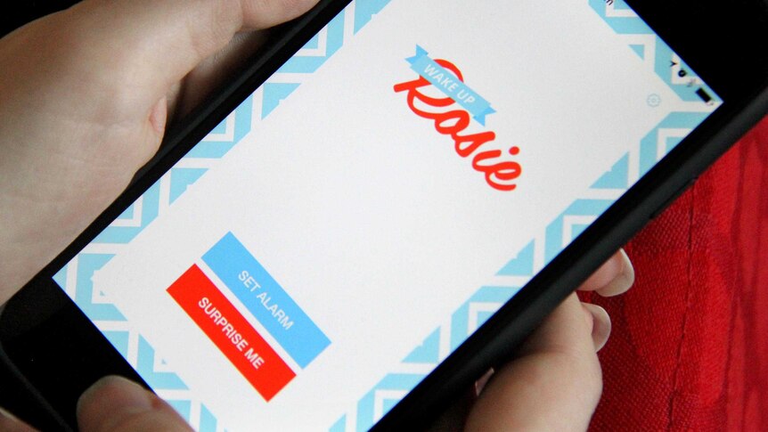 A young woman holds a smart phone showing the Wake Up Rosie app.