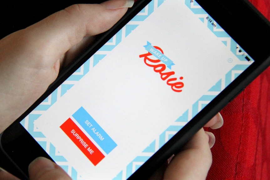 A young woman holds a smart phone showing the Wake Up Rosie app.