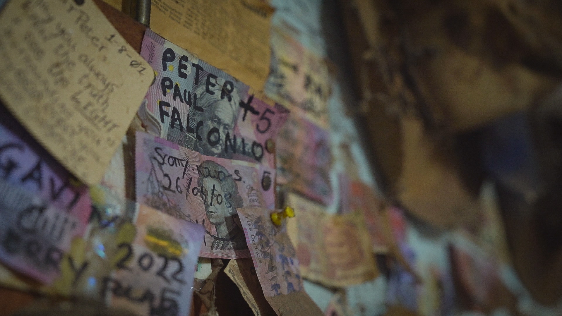 An Australian five dollar note stuck to a wall with other notes and papers reading PETER + PAUL FALCONIO