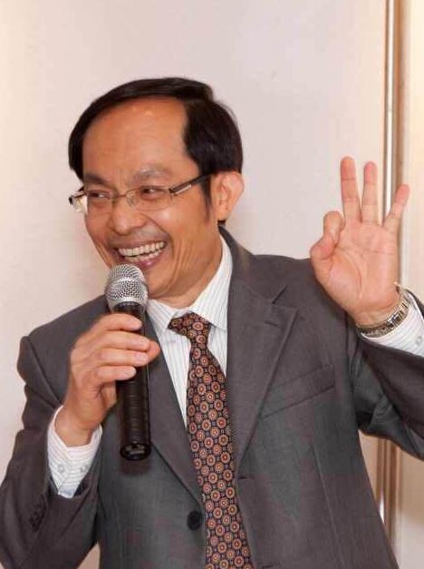 Chongyi Feng, wearing a suit, smiles and raises his hand as he speaks into a microphone.