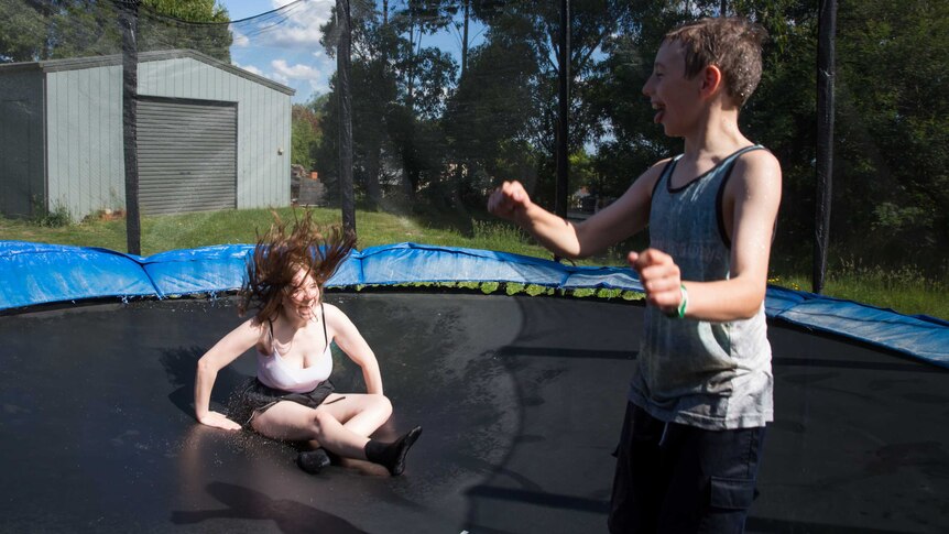 A teenage girl and younger boy laugh as they jump on a trampoline in a bushy backyard.