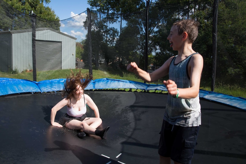 A teenage girl and younger boy laugh as they jump on a trampoline in a bushy backyard.