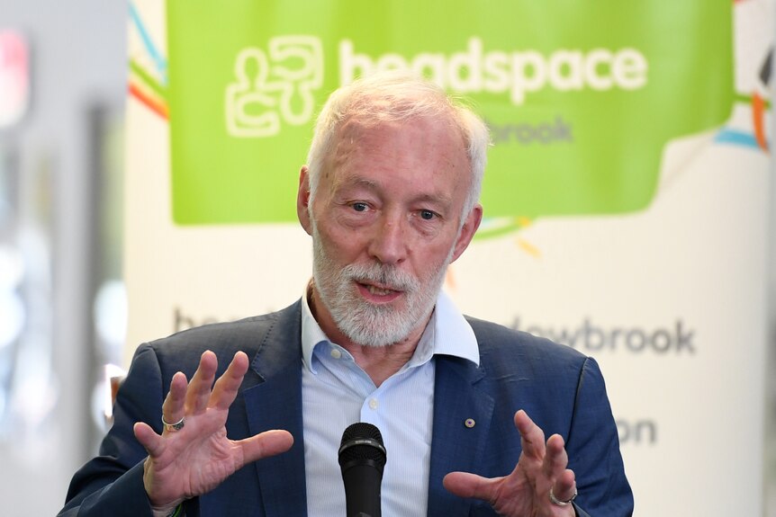 A man with grey hair and a grey beard gestures while speaking at the lectern.