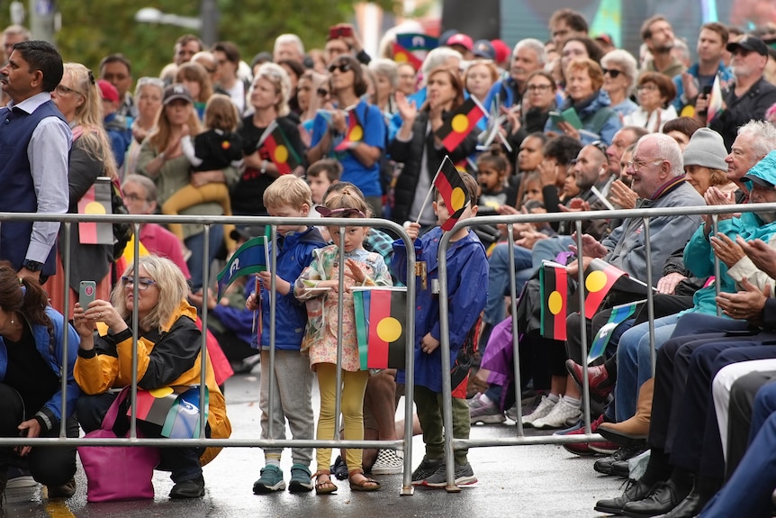 A crowd, some standing, some sitting, some waving Aboriginal and Torres Strait Islander flags behind a gate