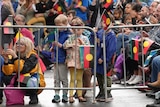 A crowd, some standing, some sitting, some waving Aboriginal and Torres Strait Islander flags behind a gate
