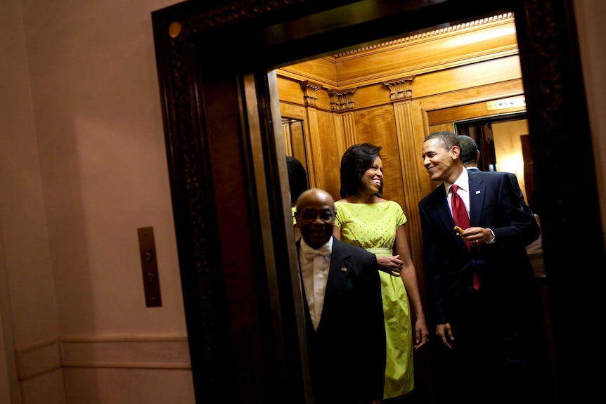 A short black man with glasses stands at front of an antique elevator with a tall black couple.