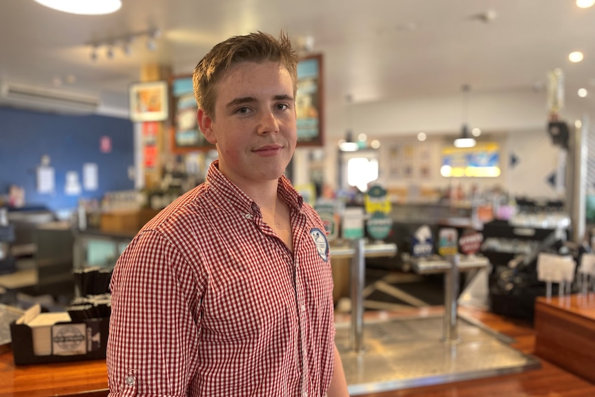 A young man in a red and white checked shirt stands at a bar.