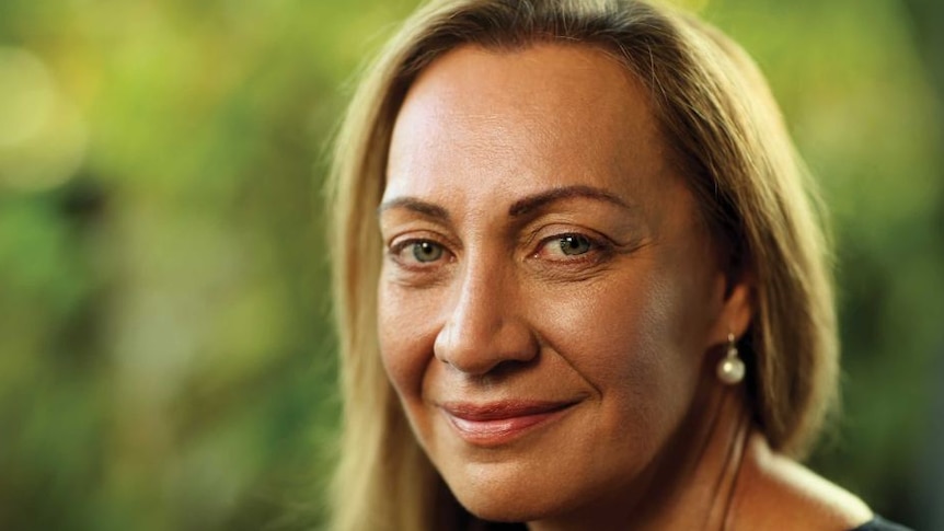 A woman with a faint smile, with her head turned towards the camera. She has brown eyes, blonde hair and pearl earrings