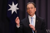 A man in a suit and tie speaks at a press conference in front of the Australian flag.