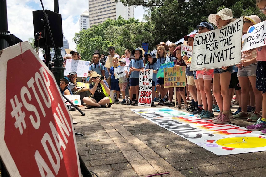 Students stand holding signs at the front of a large rally calling for action on climate change.