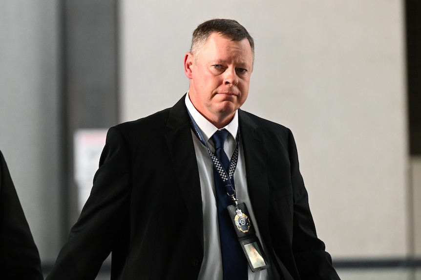 a police officer dressed in a suit with a badge on a lanyard around his neck walking out of a courthouse looking sombre