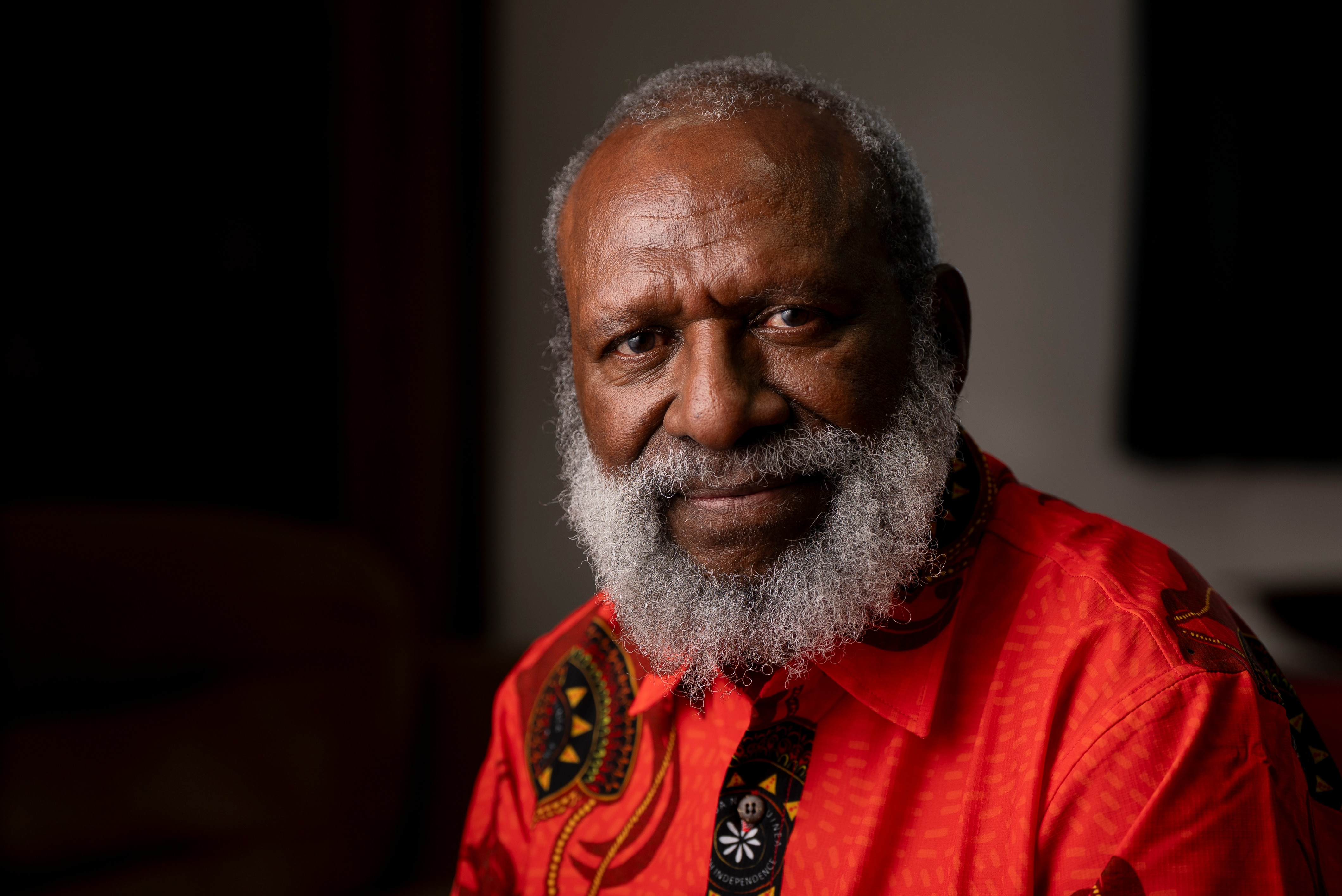 A man with a light grey beard wears a red shirt while looking neutrally at the camera in a darkened room