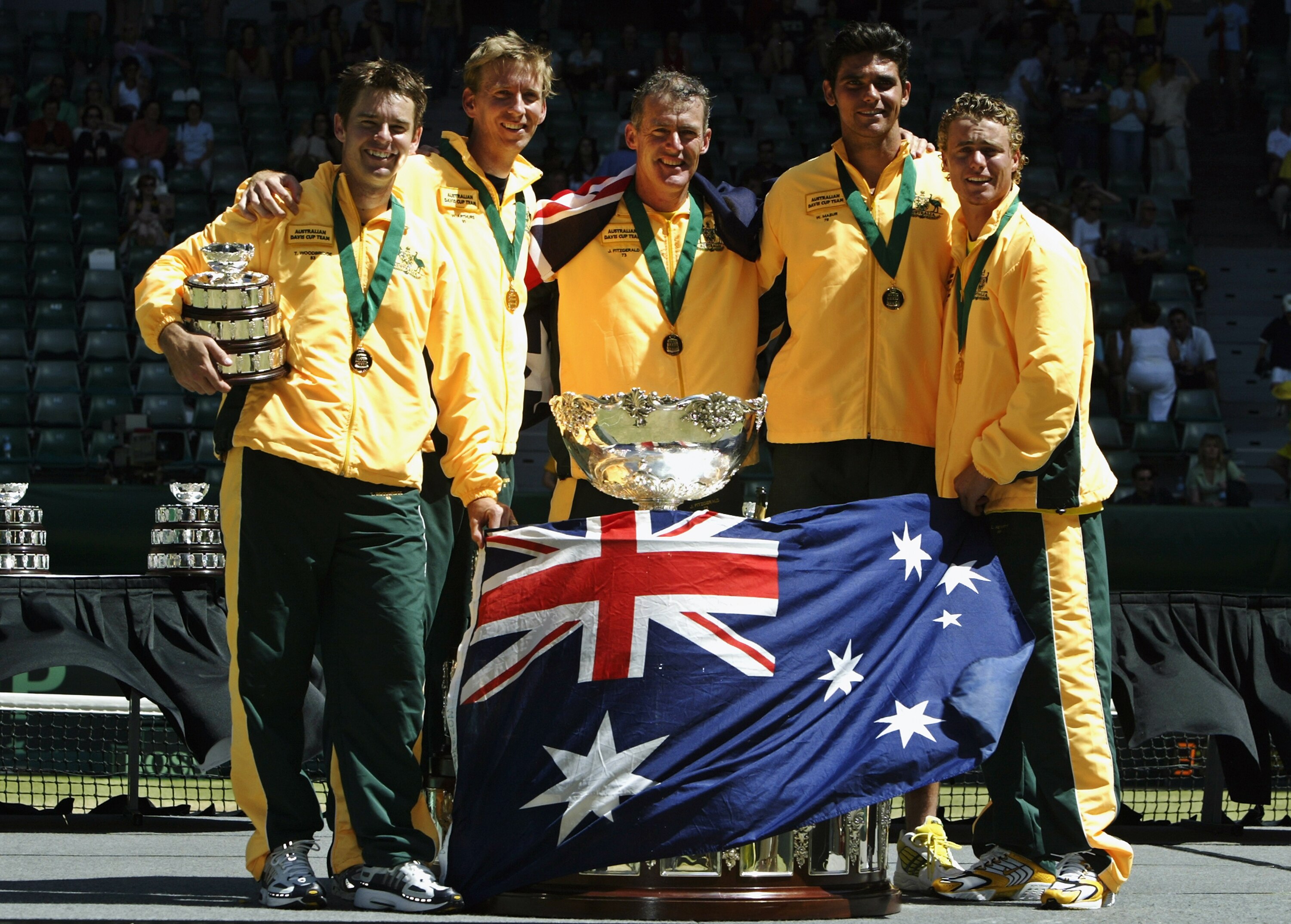 An Australian national tennis team stands next to the Davis Cup which is draped in the Australian flag.