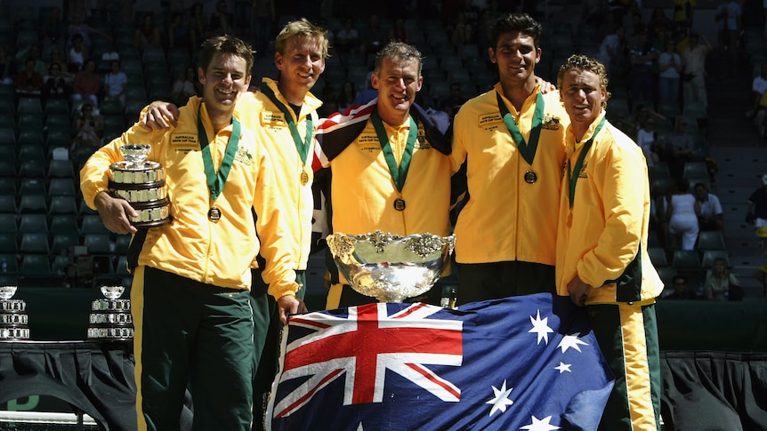 Une équipe nationale de tennis australienne se tient à côté de la Coupe Davis qui est drapée du drapeau australien.