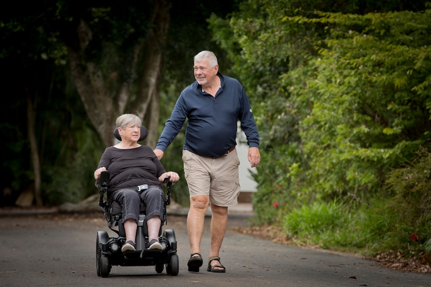 An older couple; a woman is in a motorised scooter, a man is next to her and helping her along