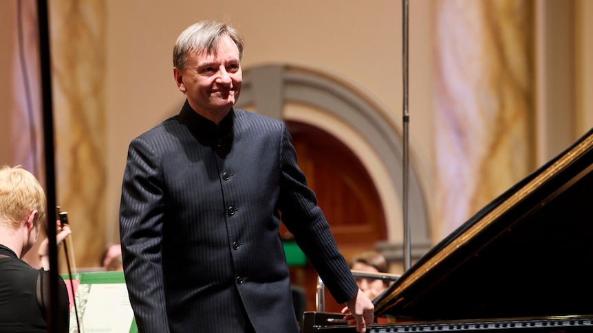 Stephen Hough is dressed in plain black and standing at the piano, smiling.