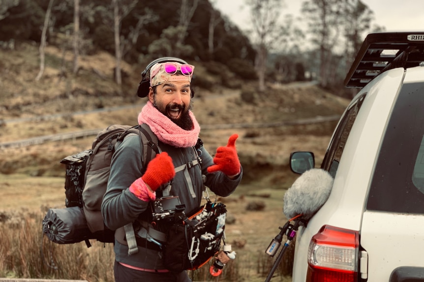 Man wearing pink hat, glasses, scarf and gloves and carrying sound recording gear. 