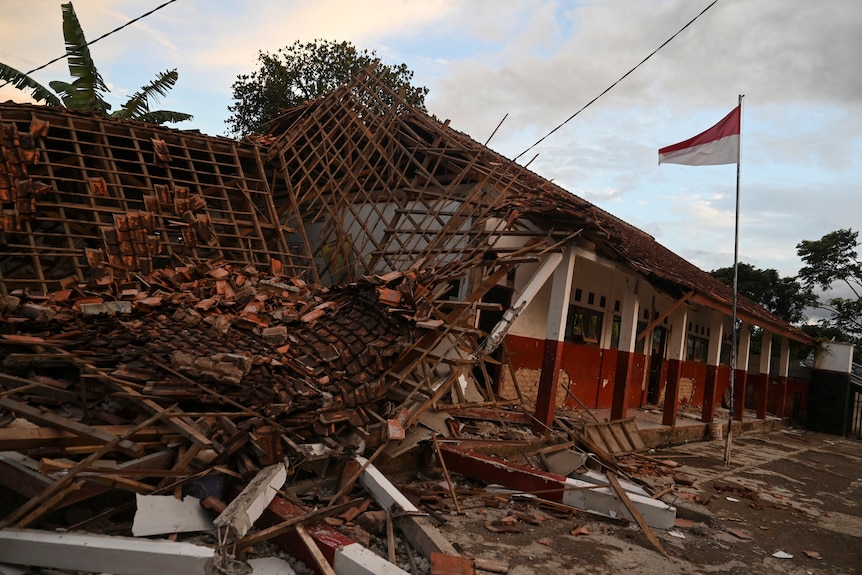 A collapsed building, with rafters, iron grills on the ground, the front red and white portion is still standing, flag in front.