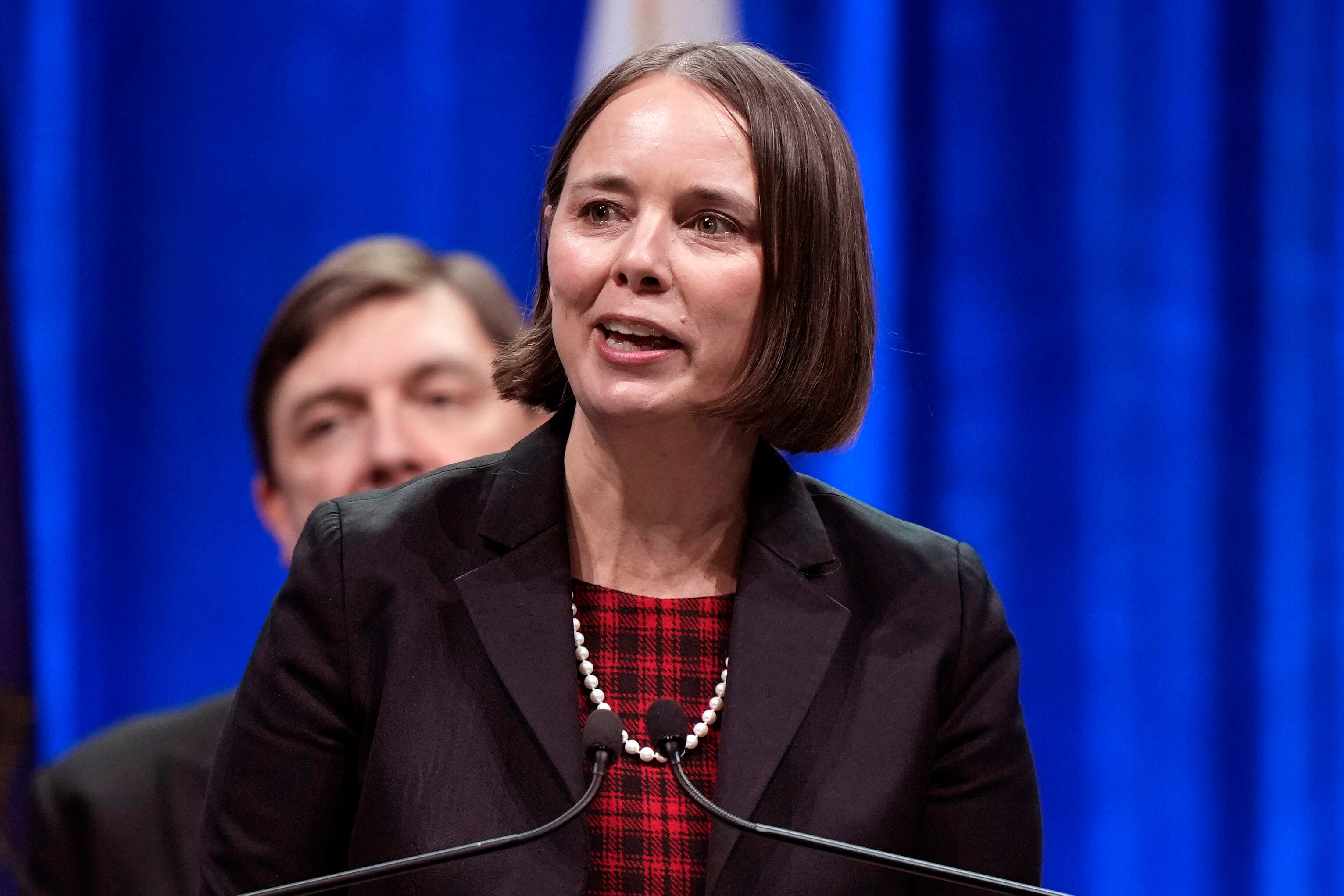 A close up of a middle-aged white woman with straight hair to her chin speaking at a microphoned lecturn. A man behind her