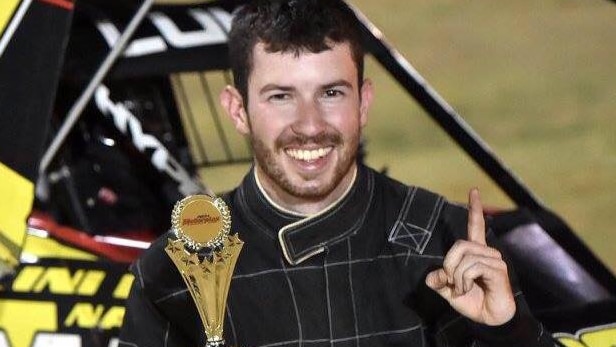 A smiling young man in a racing driver's jumpsuit holds a trophy and raises his finger in triumph.