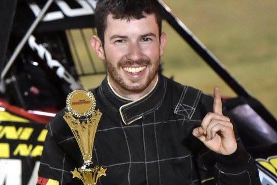 A smiling young man in a racing driver's jumpsuit holds a trophy and raises his finger in triumph.