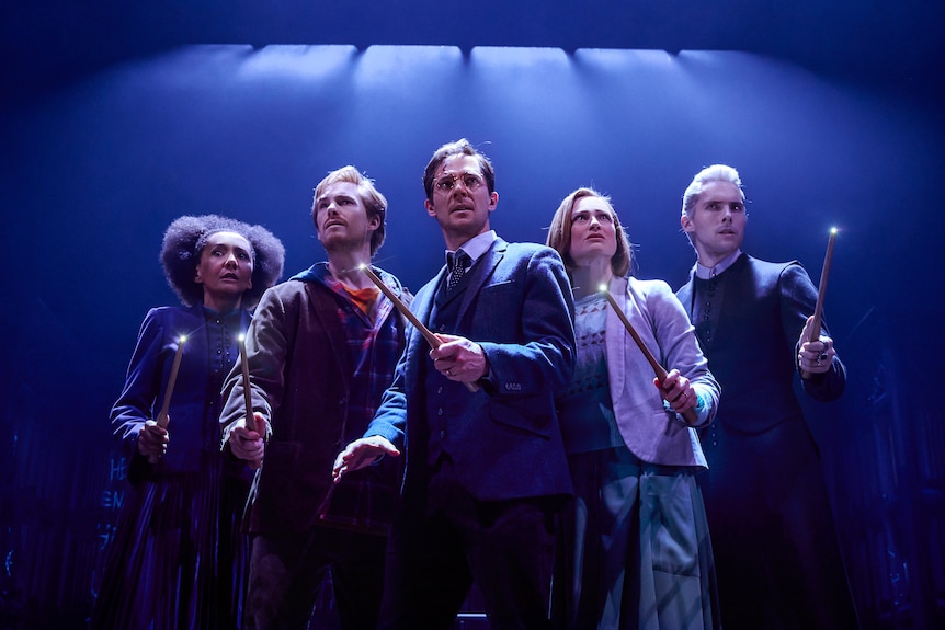 Five people stand in a row on a stage. They are all looking pensive and holding wands with lit tips.
