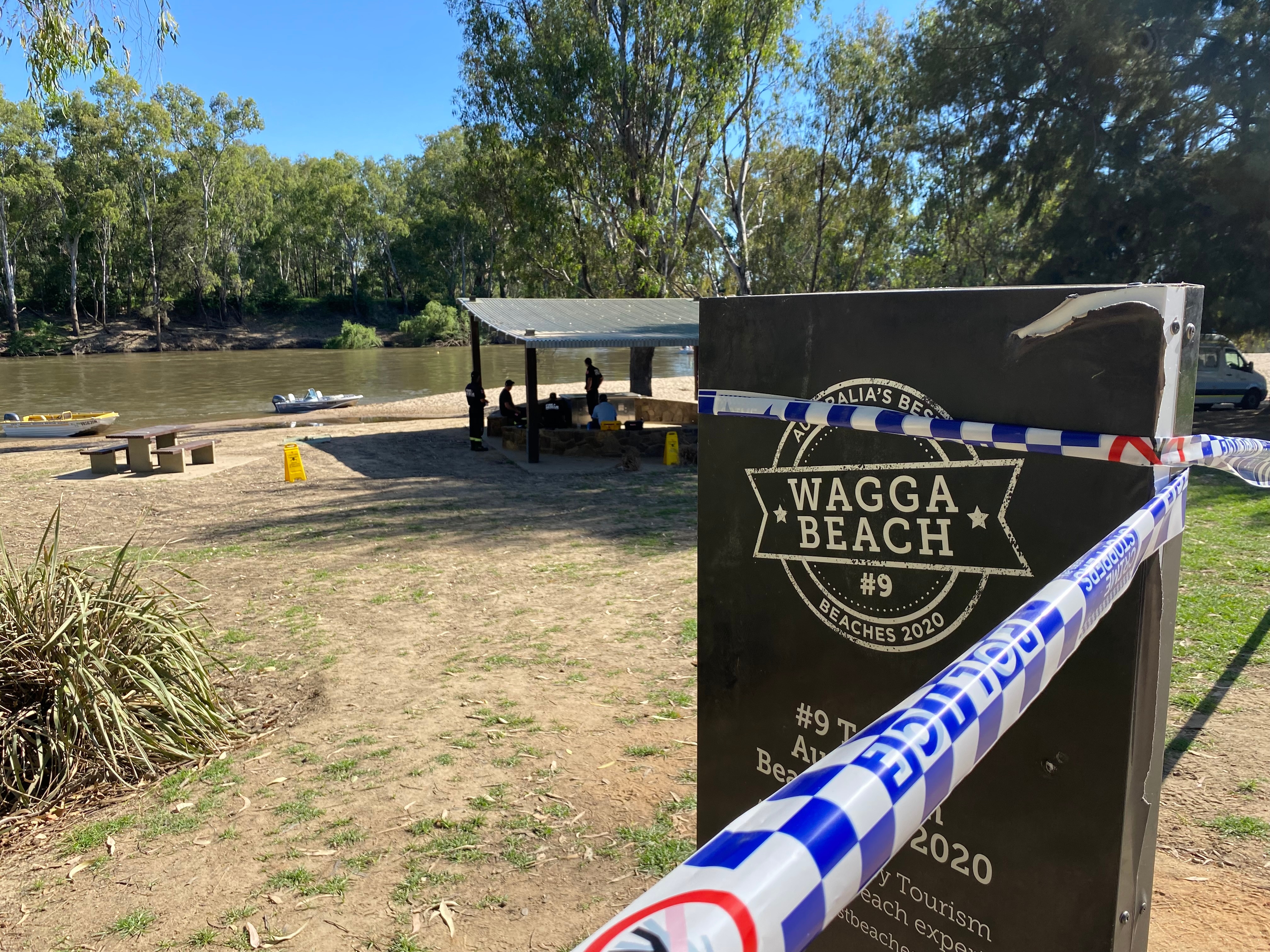 A sign saying 'Wagga Beach' with police tape around it and boats in the river behind it.