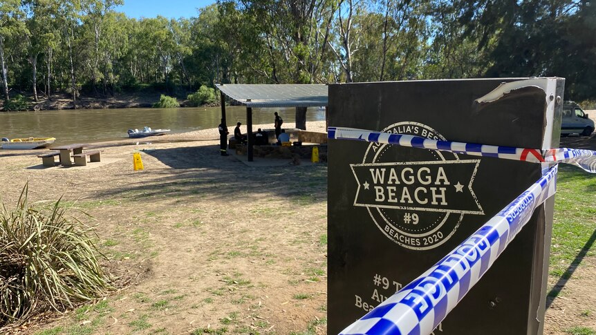 A sign saying 'Wagga Beach' with police tape around it and boats in the river behind it.