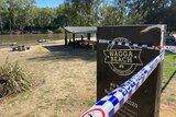 A sign saying 'Wagga Beach' with police tape around it and boats in the river behind it.